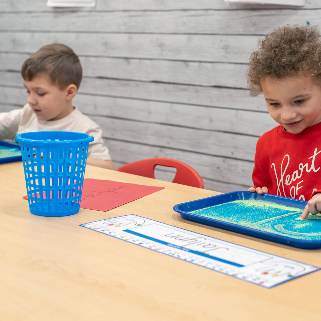 “Two young children using trays of colored sand for a sensory-learning activity in a classroom.”
